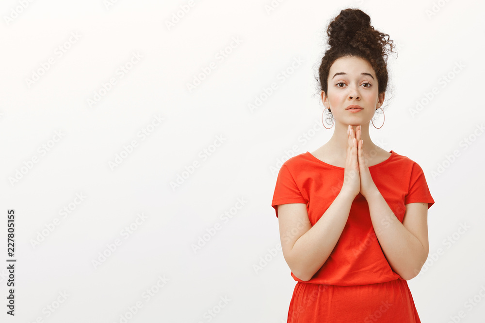 Indoor shot of positive cute girlfriend in red dress with curly hair combed in bun, holding hands in pray above chin and gazing with tender expression at camera while asking help or favour, begging Indoor shot of positive cute girlfriend in red dress with curly hair combed in bun, holding hands in pray above chin and gazing with tender expression at camera while asking help or favour, begging