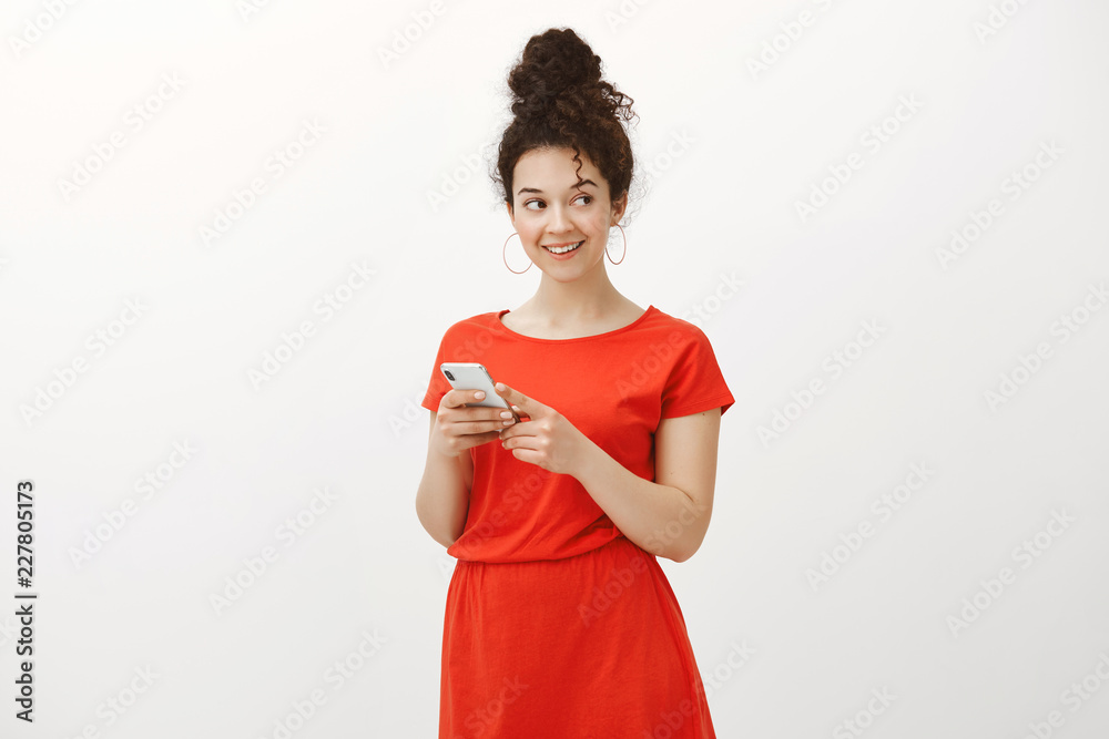 Intrigued friendly european woman in stylish red dress, holding brand new smartphone and gazing right with satisfied flirty smile, discussing hot guy with friend via messages over gray wall Intrigued friendly european woman in stylish red dress, holding brand new smartphone and gazing right with satisfied flirty smile, discussing hot guy with friend via messages over gray wall