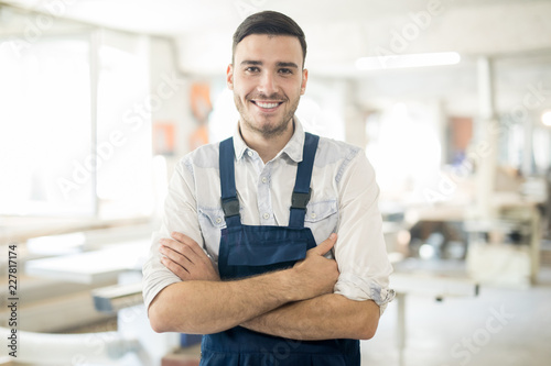 Young successful worker in overalls crossing his arms on chest and looking at you in workshop photo