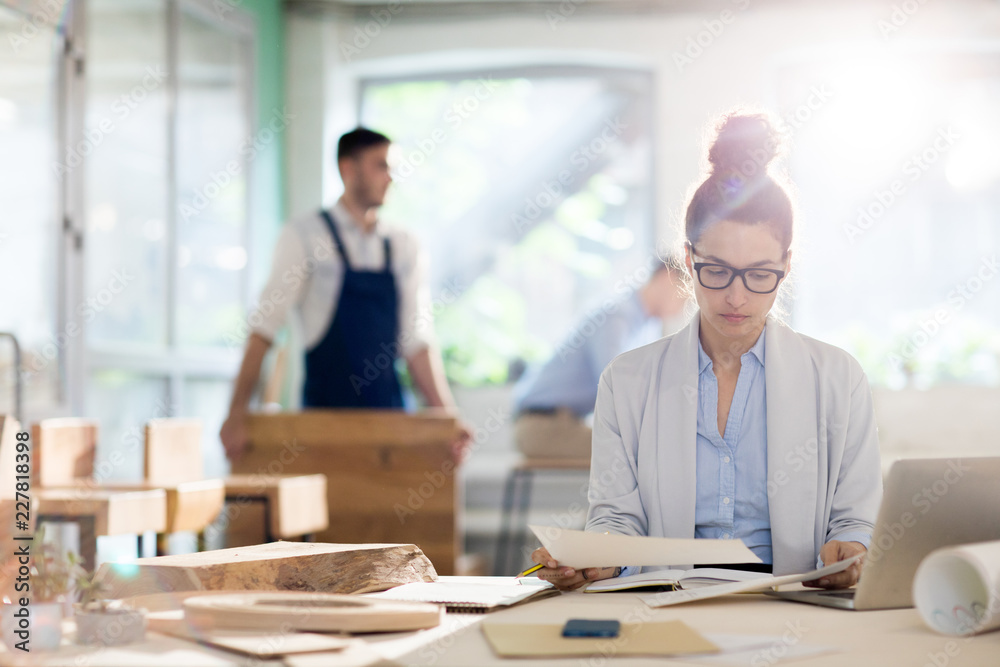 Young analyst looking through working papers or sketches while sitting by desk with colleagues on background Young analyst looking through working papers or sketches while sitting by desk with colleagues on background