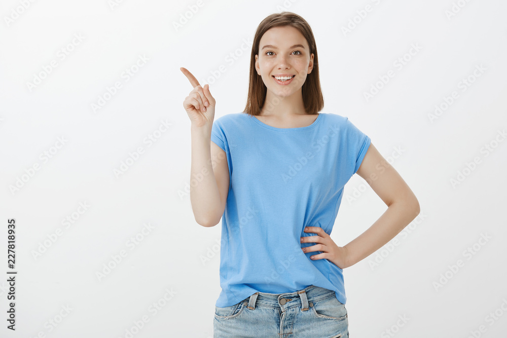 Look, awesome copy space. Positive happy woman in blue blouse, holding hand on hip and pointing at upper left corner with index finger, smiling broadly, being satisfied and pleased new cafe Look, awesome copy space. Positive happy woman in blue blouse, holding hand on hip and pointing at upper left corner with index finger, smiling broadly, being satisfied and pleased new cafe