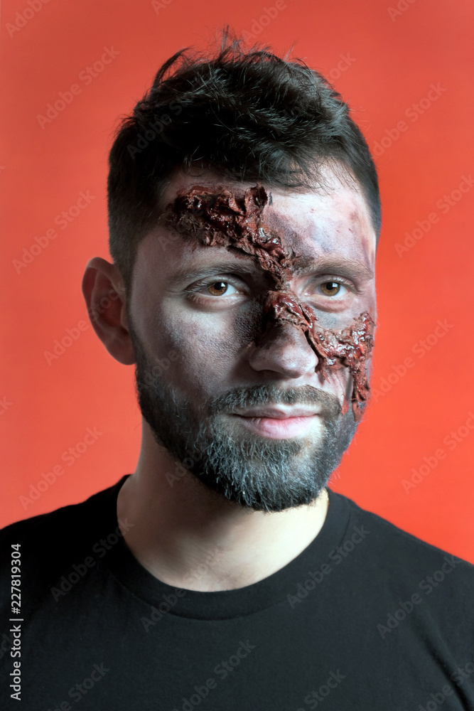 Obraz premium The guy in the black t-shirt with makeup in the style of Halloween. Vertical portrait of a young man with ragged facial wounds. Festive party. Red background.