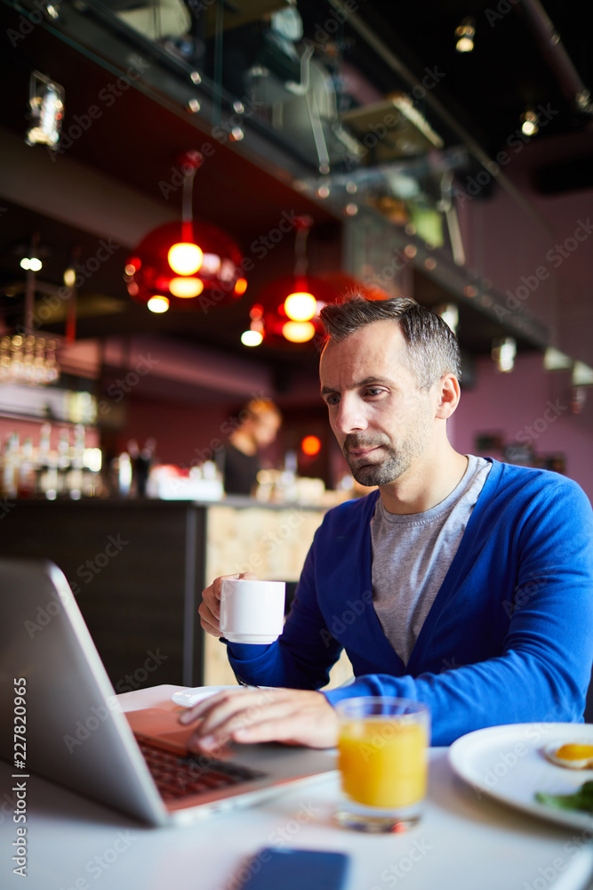 Modern businessman having tea or coffee while sitting by table in front of laptop and networking
