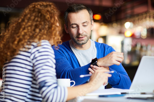 Young businessman showing time on smartwatch to his colleague while sitting in cafe before briefing or seminar