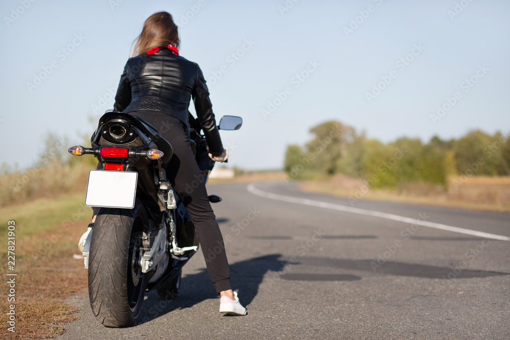 Obraz premium Back view of stylish biker on motorbike, covers long destination, rides in countryside in open road, enjoys time for relax, likes her transport, feels freedom and independence. Outdoor activity