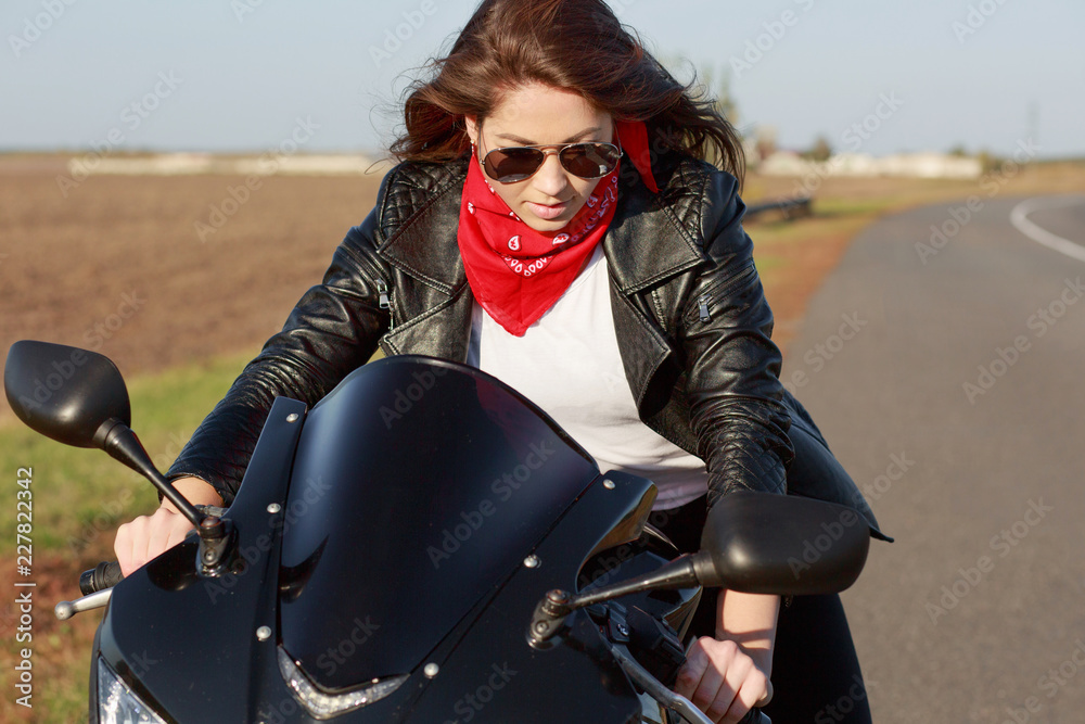 Good looking young woman in stylish black leather jacket, red bandana and sunglasses, rides motorbike, poses on road with no people and transport, enjoys high speed, spends leisure time outdoor