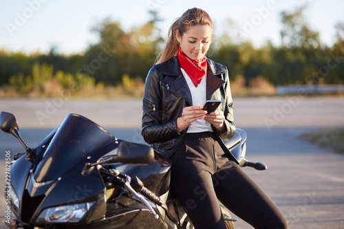 Photo of female driver holds smart phone in hands, surfes bikers blog in internet, wears stylish clothes, leans at black fast motorbike. Beautiful biker plans rout, uses map on modern mobile phone