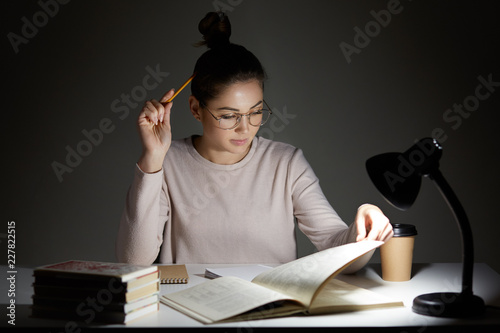 Busy student absorbed in reading, wears round transparent glasses for good vision, uses reading lamp, sits at workplace, works in darkness, holds pencil, has clever look, prepares for final exam photo