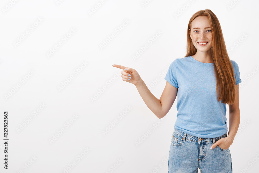 Cute friendly girl showing granny way to store. Portrait of charming redhead young female with freckles holding hands in pocket and pointing left with joyful polite smile over gray background Cute friendly girl showing granny way to store. Portrait of charming redhead young female with freckles holding hands in pocket and pointing left with joyful polite smile over gray background
