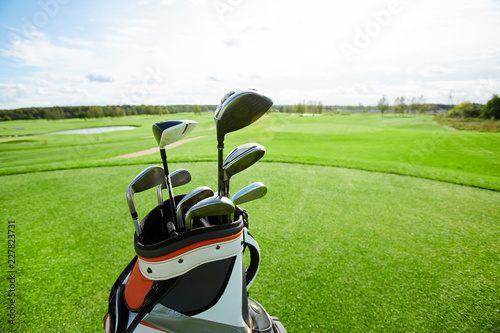 Bag with bunch of golf clubs and vast green field for playing golf on background