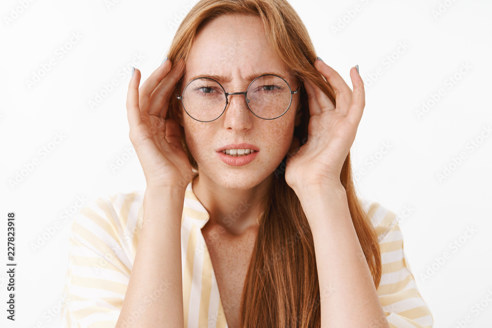 Concerned clueless and perplexed good-looking stylish female student in glasses frowning and squinting holding hands on temples of head being troubled or suffering headache over white wall
