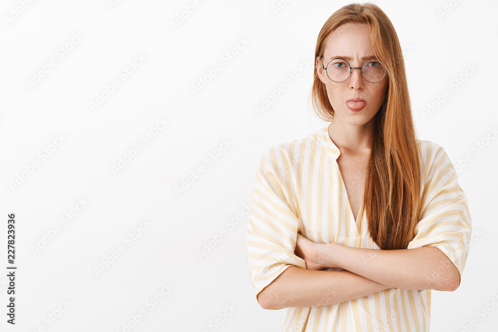 Waist-up shot of moody displeased and offended sad redhead girl with freckles holding hands crossed on chest frowning and showing tongue feeling insulted over gray background Waist-up shot of moody displeased and offended sad redhead girl with freckles holding hands crossed on chest frowning and showing tongue feeling insulted over gray background