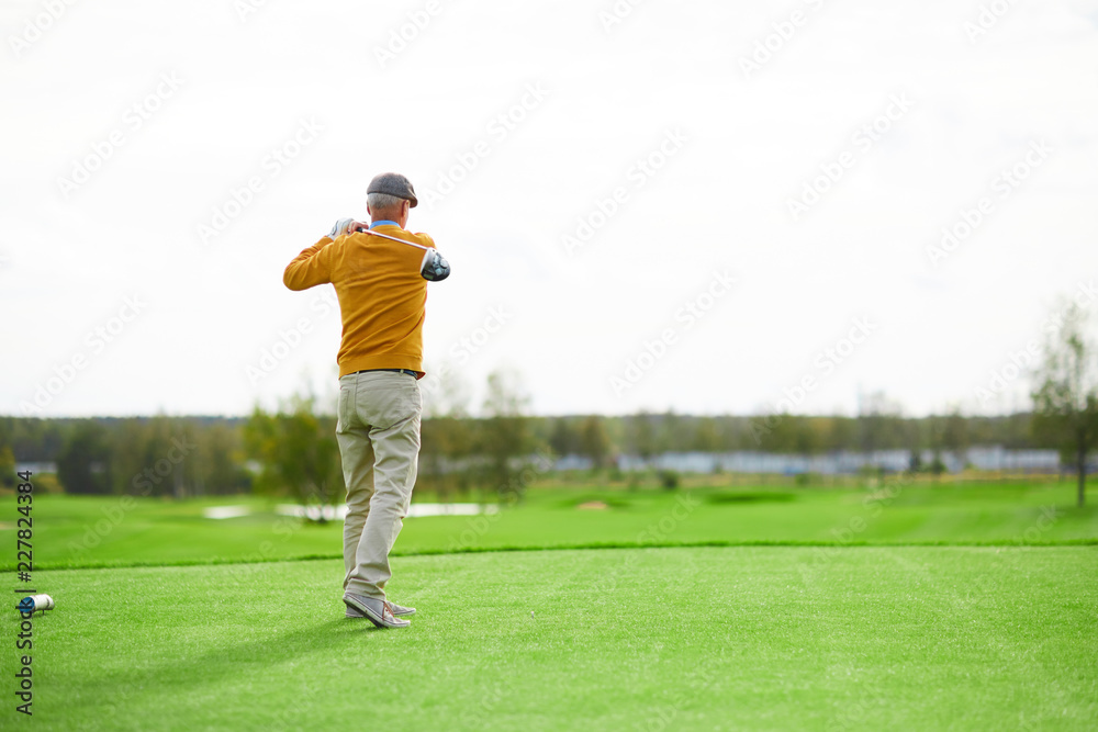 Back view of senior man in cap, yellow pullover and beige pants hit ball with golf club on green field