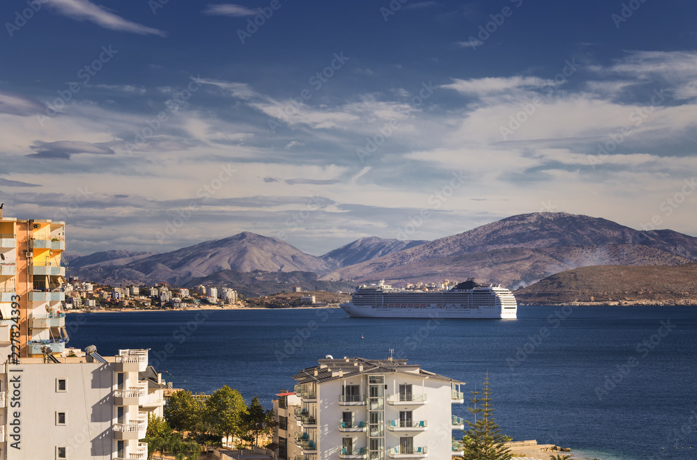 Obraz premium Big cruise ship sails to the sea port of Saranda, Albania. Albanian mountains in the background. Sunset, cloudy sky.