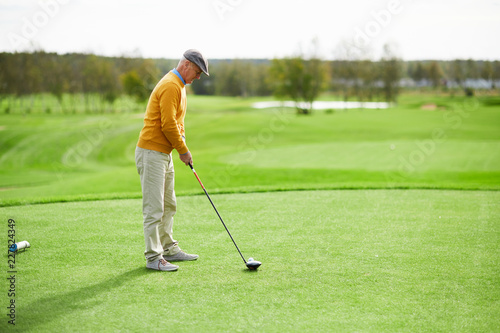 Senior casual man standing on green lawn and keeping golf club close to ball on grass