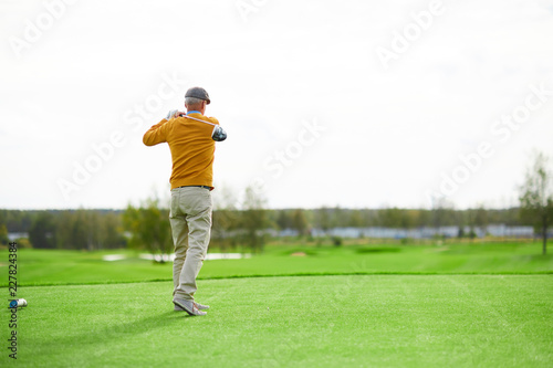 Back view of senior man in cap, yellow pullover and beige pants hit ball with golf club on green field
