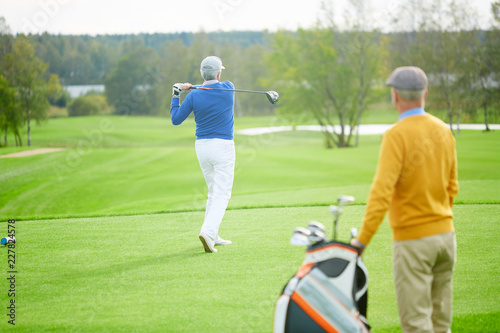 Aged man hit golf ball during leisure game while his buddy with bag for clubs standing near by