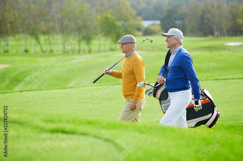 Two senior sportsmen with golf clubs walking along vast green field while hurrying for leisure game