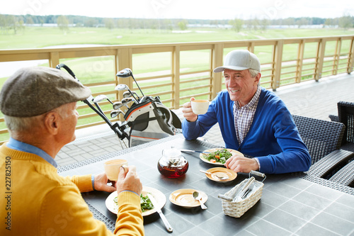 Two mature golf players sitting by table in front of one another, having salad and tea and discussing the last game
