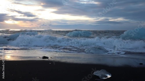 Icebergs and pieces of blue and white ice are rolling on the waves. Waves breaking on the ice. Surf on the beach at sunrise. Iceland