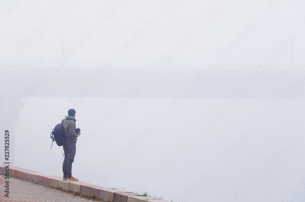 Man photographer looking at bridge holding camera in his hands on foggy morning. Rear view. Copy space.