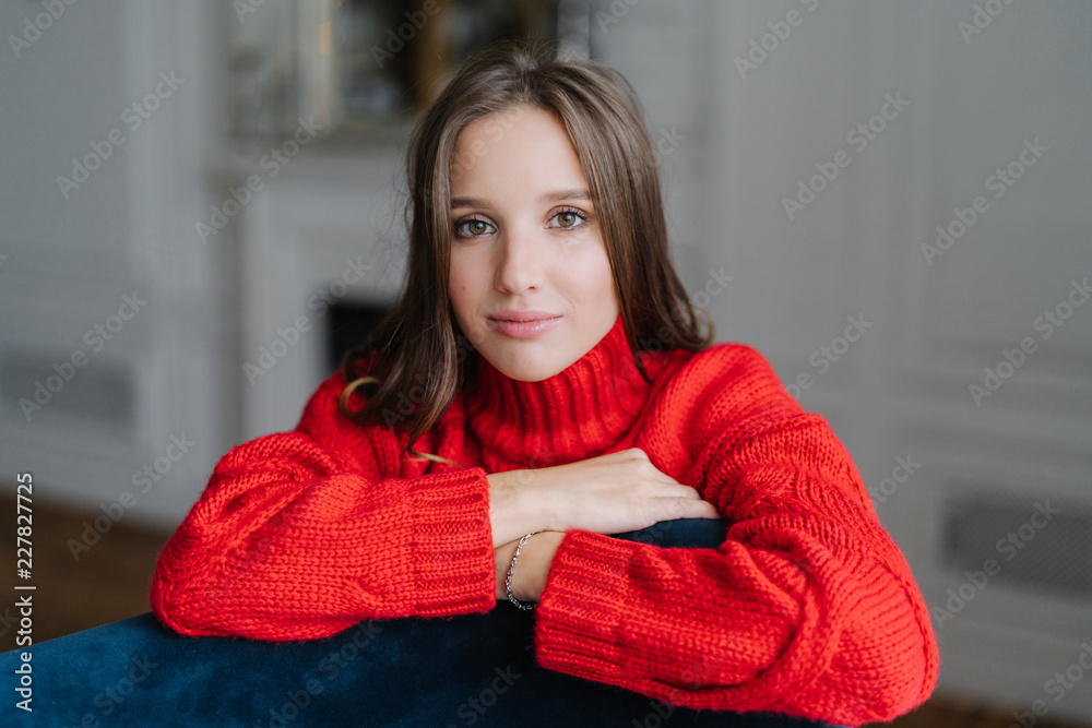Calm pretty young Caucasian female dressed in warm knitted pullover, leans on armchair, looks with dark eyes directly at camera, poses against domestic interior indoor. People and beauty concept. Calm pretty young Caucasian female dressed in warm knitted pullover, leans on armchair, looks with dark eyes directly at camera, poses against domestic interior indoor. People and beauty concept.