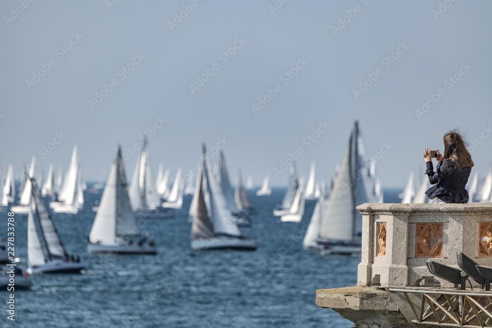 Fototapeta premium Donna giovane che fotografa con cellulare una regata velica con tante barche a vela