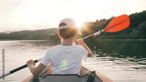 Slow motion a happy boy with her dog Jack Russell breed paddling on an inflatable kayak on the water of a large mountain lake on the background of a beautiful orange sunset. Family sports holiday