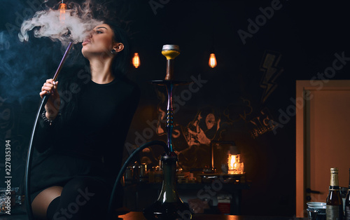 Sexy brunette girl in seductive black clothes smokes a hookah while sitting on counter in a nightclub or bar.