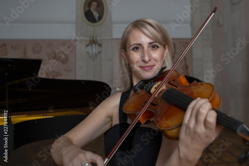 Young beautiful woman with wavy blonde hair playing viola, holding bow hovering over instrument on her shoulder and smiling, looking at camera