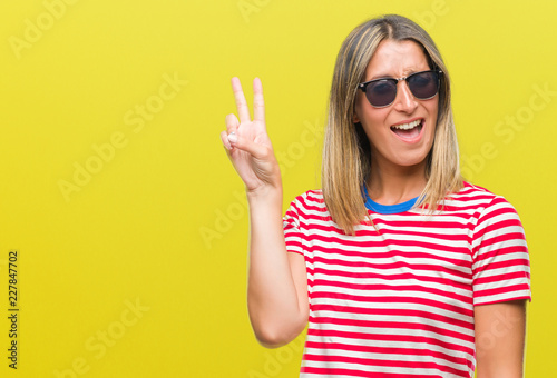 Young beautiful woman wearing sunglasses over isolated background smiling with happy face winking at the camera doing victory sign. Number two.