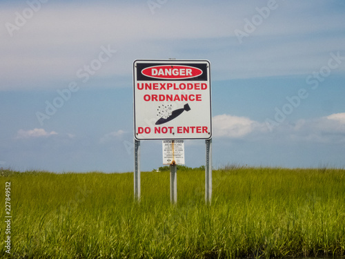 Danger unexploded ordnance do not enter  posted sign above green sea grass in front of a blue sky wide view