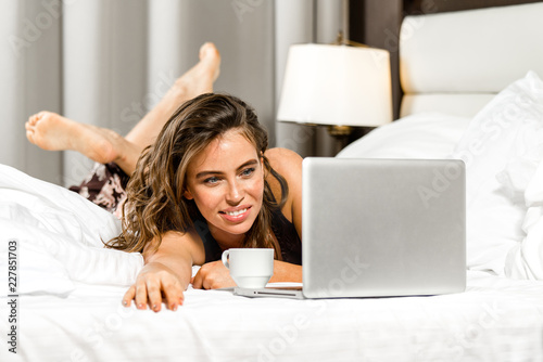 attractive young caucasian cmiling cheerful woman at home in bed with cup of tea or coffee with notebook laptop looking at notebook working at home