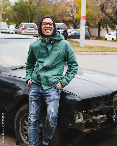 Man in glasses and jacket is smiling at the wrecked car photo