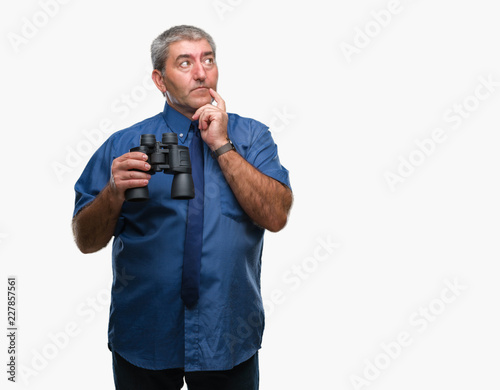 Handsome senior man looking through binoculars over isolated background serious face thinking about question, very confused idea