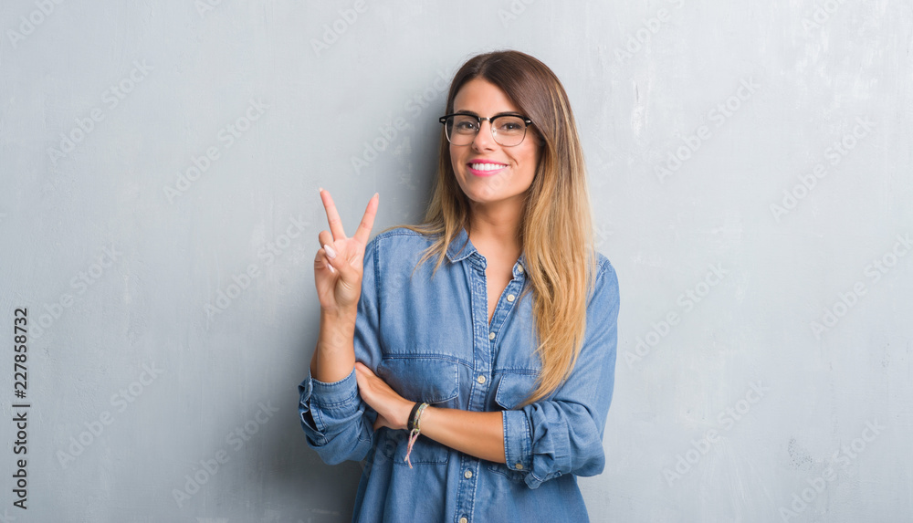 Young adult woman over grunge grey wall wearing glasses smiling with happy face winking at the camera doing victory sign. Number two. Young adult woman over grunge grey wall wearing glasses smiling with happy face winking at the camera doing victory sign. Number two.