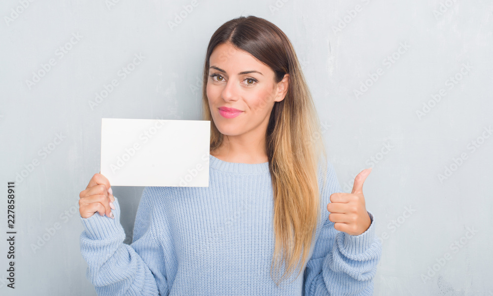 Young adult woman over grey grunge wall holding blank card happy with big smile doing ok sign, thumb up with fingers, excellent sign Young adult woman over grey grunge wall holding blank card happy with big smile doing ok sign, thumb up with fingers, excellent sign