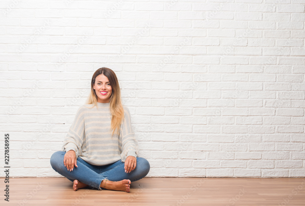 Young adult woman sitting on the floor over white brick wall at home with a happy and cool smile on face. Lucky person. Young adult woman sitting on the floor over white brick wall at home with a happy and cool smile on face. Lucky person.