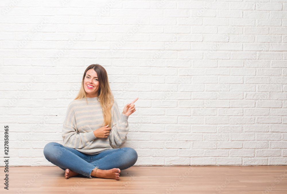 Young adult woman sitting on the floor over white brick wall at home with a big smile on face, pointing with hand and finger to the side looking at the camera. Young adult woman sitting on the floor over white brick wall at home with a big smile on face, pointing with hand and finger to the side looking at the camera.