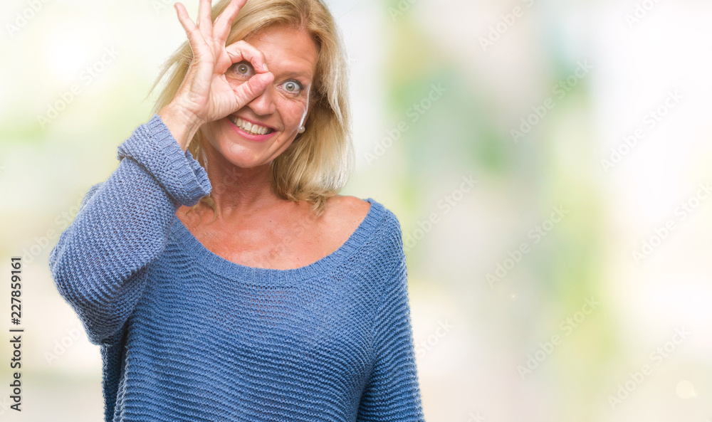 Middle age blonde woman wearing winter sweater over isolated background doing ok gesture with hand smiling, eye looking through fingers with happy face.