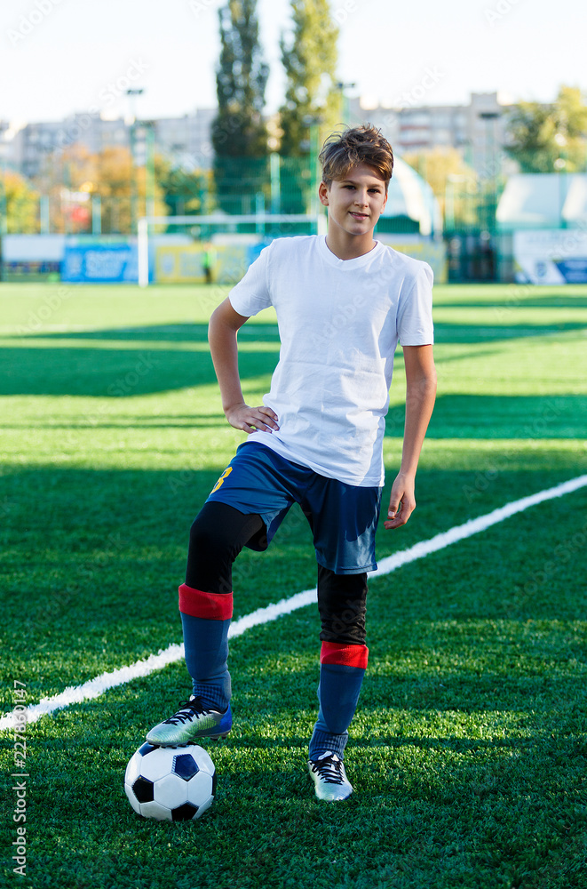 cute young boy in white blue sportswear stands next to classical black and white football ball on the stadium field. Soccer game, training, hobby concept. 