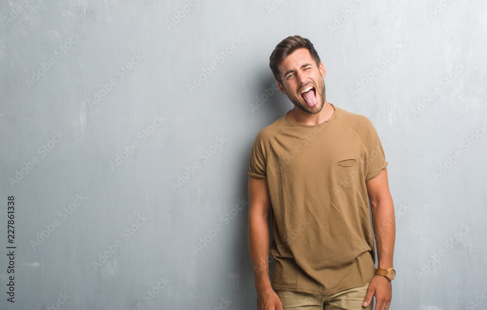 Handsome young man over grey grunge wall sticking tongue out happy with funny expression. Emotion concept.