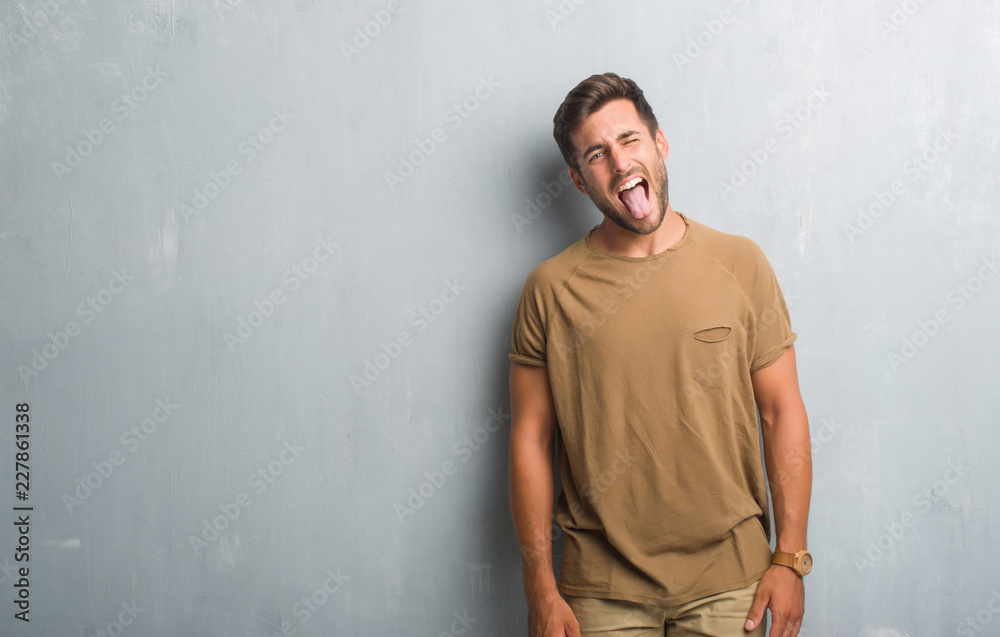 Handsome young man over grey grunge wall sticking tongue out happy with funny expression. Emotion concept. Handsome young man over grey grunge wall sticking tongue out happy with funny expression. Emotion concept.