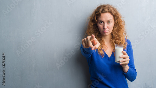 Young redhead woman drinking glass of milk pointing with finger to the camera and to you, hand sign, positive and confident gesture from the front