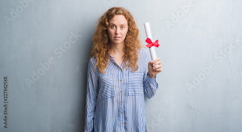 Young redhead business woman over grey grunge wall holding diploma with a confident expression on smart face thinking serious