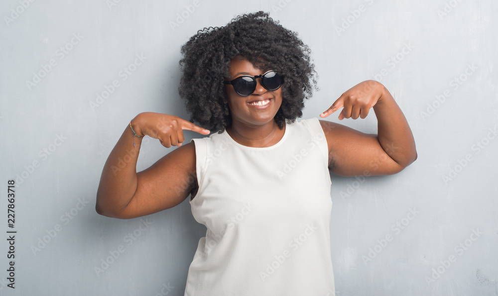 Young african american plus size woman over grey grunge wall wearing fashion sunglasses looking confident with smile on face, pointing oneself with fingers proud and happy. Young african american plus size woman over grey grunge wall wearing fashion sunglasses looking confident with smile on face, pointing oneself with fingers proud and happy.