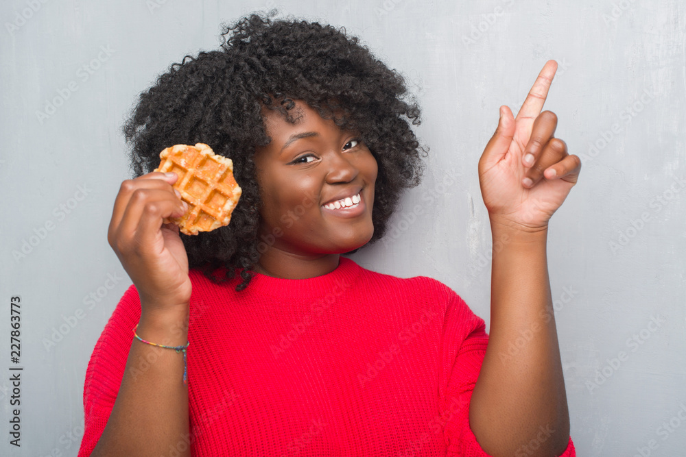 Young african american woman over grey grunge wall eating belgium waffle surprised with an idea or question pointing finger with happy face, number one Young african american woman over grey grunge wall eating belgium waffle surprised with an idea or question pointing finger with happy face, number one