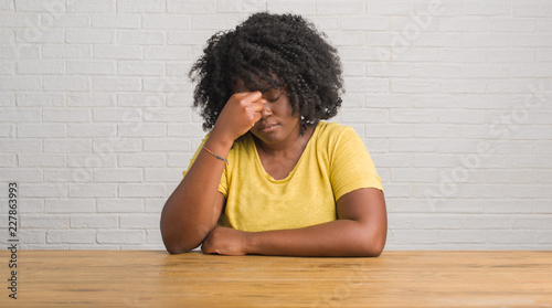 Young african american woman sitting on the table at home tired rubbing nose and eyes feeling fatigue and headache. Stress and frustration concept. photo