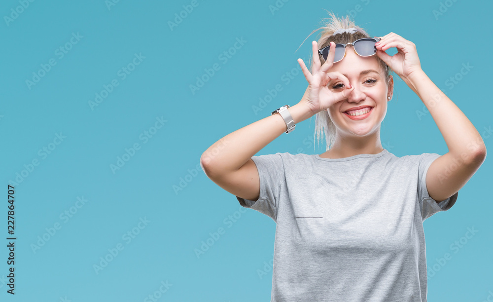 Young blonde woman wearing sunglasses over isolated background doing ok gesture with hand smiling, eye looking through fingers with happy face.