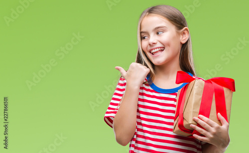 Young beautiful girl giving christmas or valentine gift over isolated background pointing and showing with thumb up to the side with happy face smiling